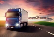 A blue truck travels along the road, with a large ship visible in the background.