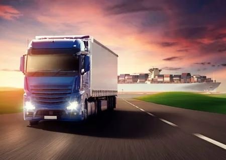 A blue truck travels along the road, with a large ship visible in the background.